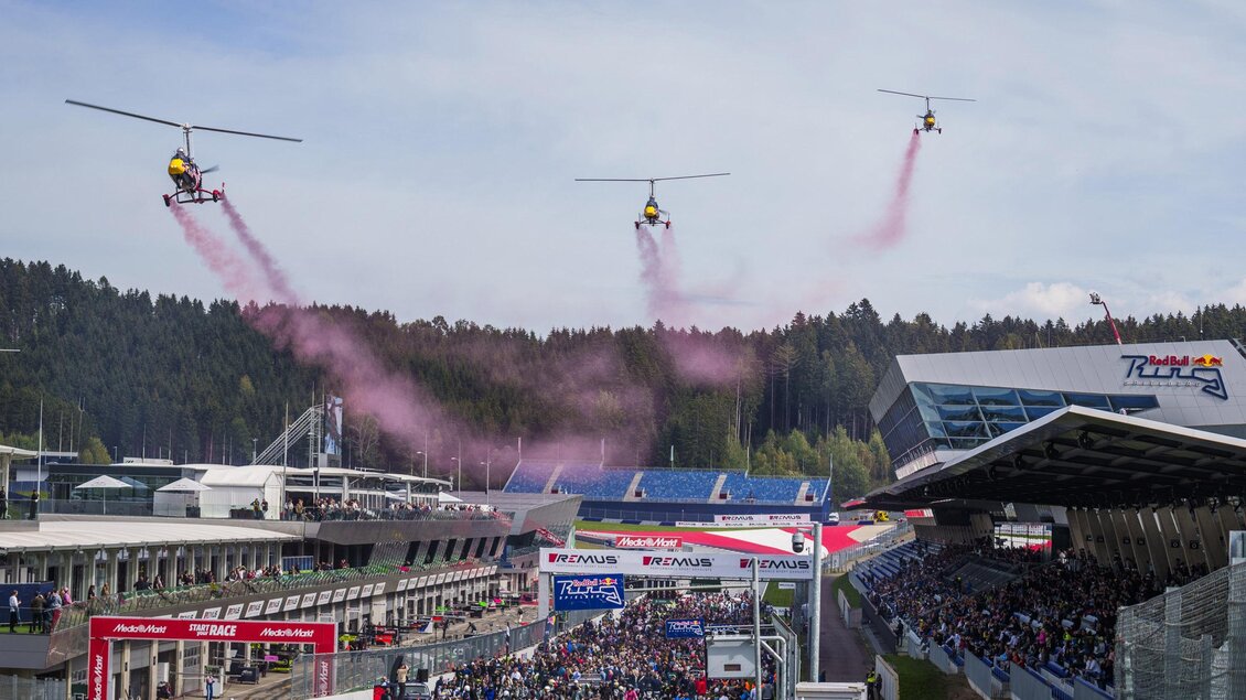 Drei Hubschrauber fliegen über eine große Menschenmenge und hinterlassen pinke Rauchspuren. Im Hintergrund ist die Rennstrecke und ein Gebäude zu sehen. | © Red Bull Ring