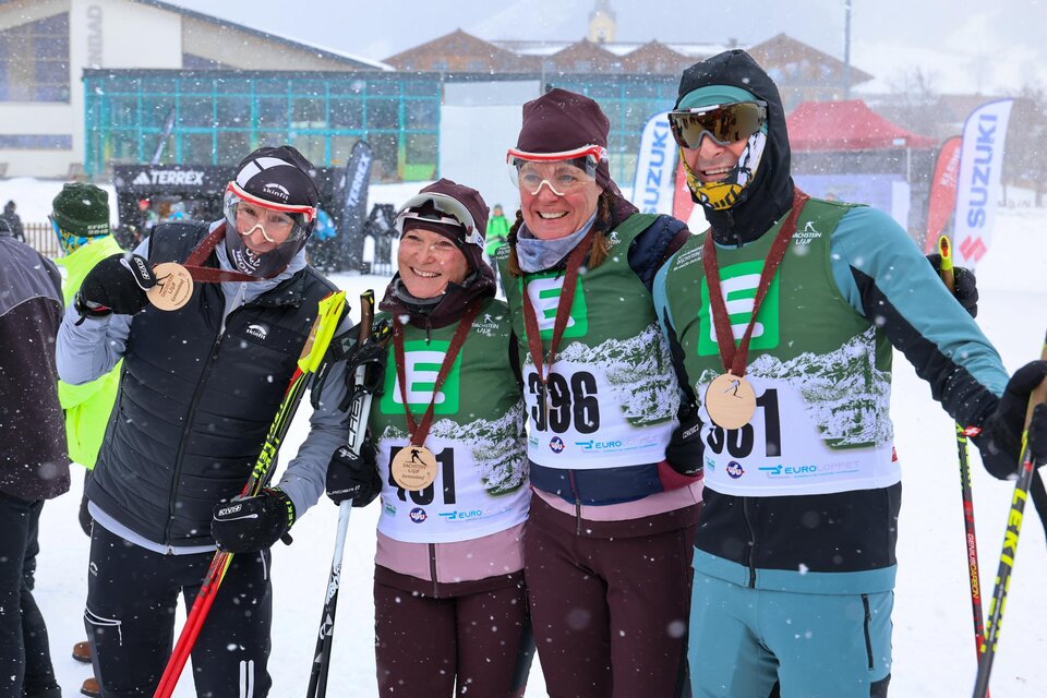 Drei Skifahrer posieren im Schnee und halten stolz ihre Medaillen. Im Hintergrund sind verschneite Berge und eine Menschenmenge zu sehen. | © Michael Simonlehner