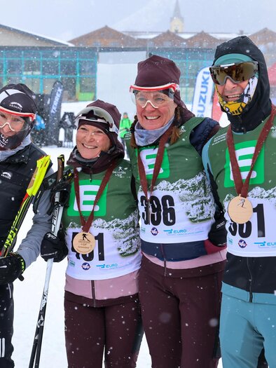 Three skiers pose in the snow, proudly holding their medals. In the background, snow-covered mountains and a crowd can be seen. | © Michael Simonlehner