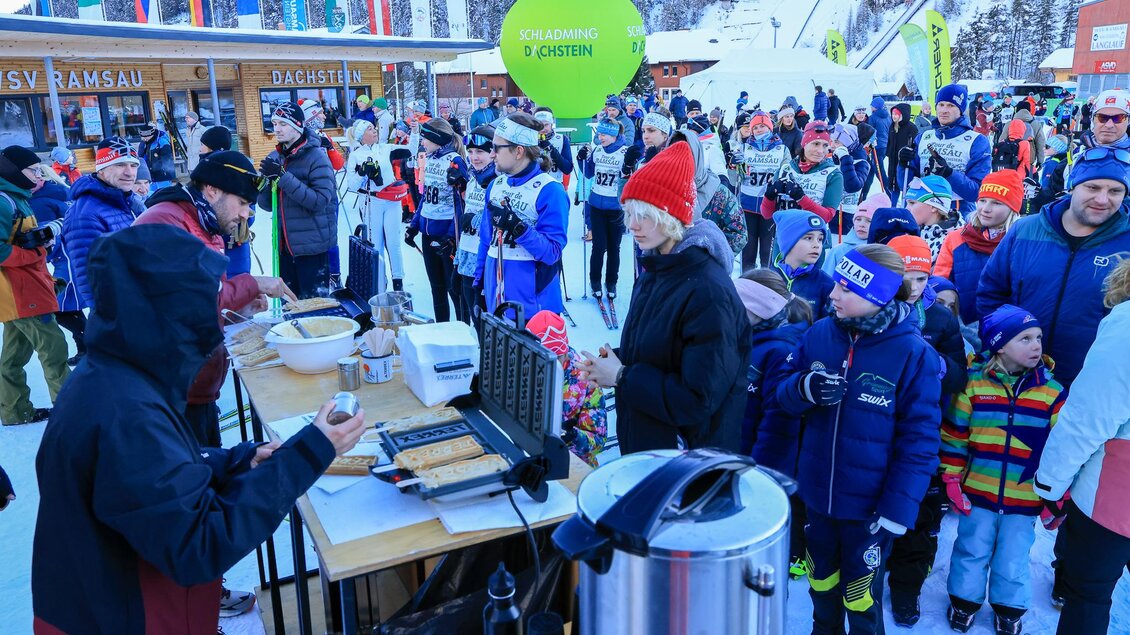 Ein lebhaftes Wintersport-Event mit vielen Teilnehmern in Skibekleidung. Im Vordergrund wird Essen und Trinken an einem Holzstand angeboten, während sich die Menschen in einer Schlange versammeln. | © Michael Simonlehner