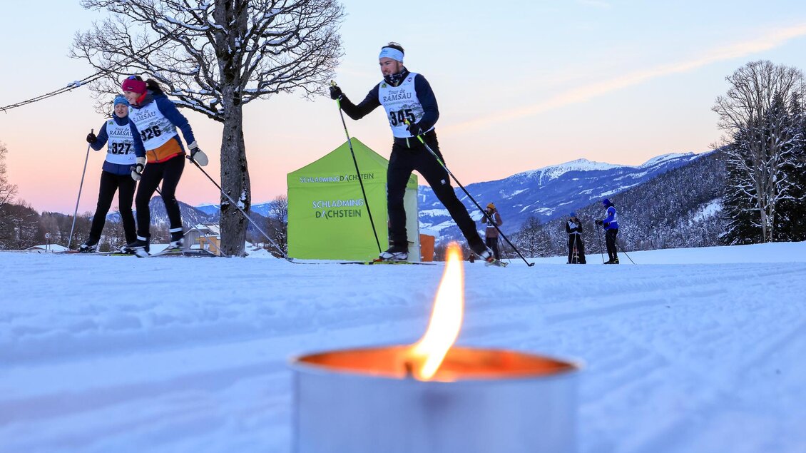 Eine Gruppe von Langläufern auf einer verschneiten Strecke bei Sonnenuntergang. Im Vordergrund brennt eine Kerze. | © Michael Simonlehner