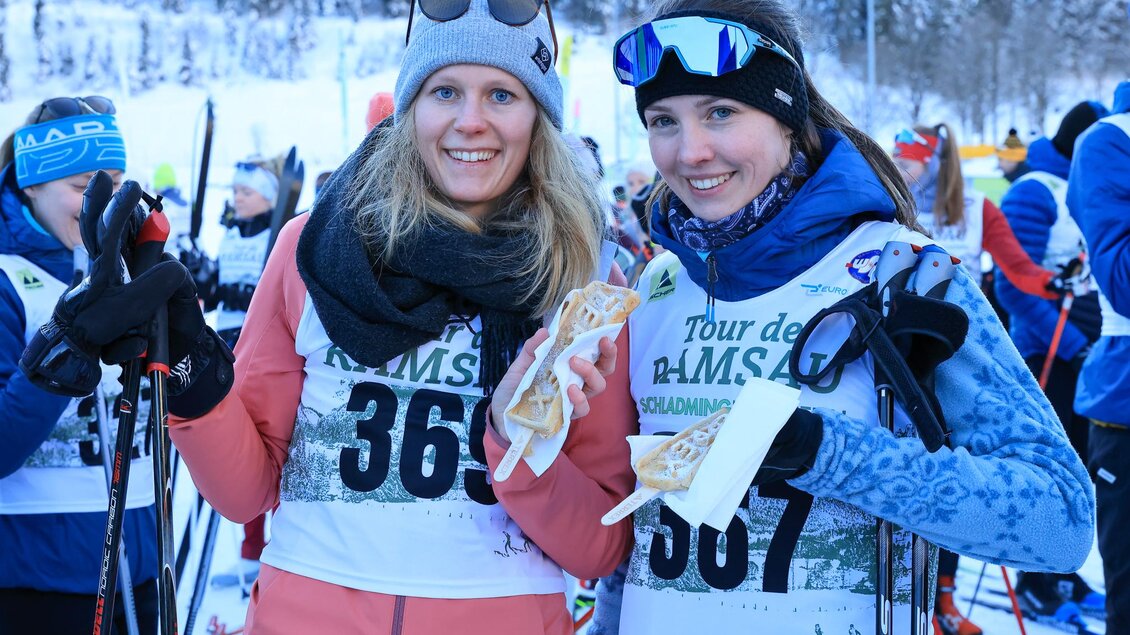 Zwei lächelnde Frauen in Skibekleidung halten Skistöcke in der Hand. Sie stehen in einer verschneiten Umgebung bei einem Skievent. | © Michael Simonlehner