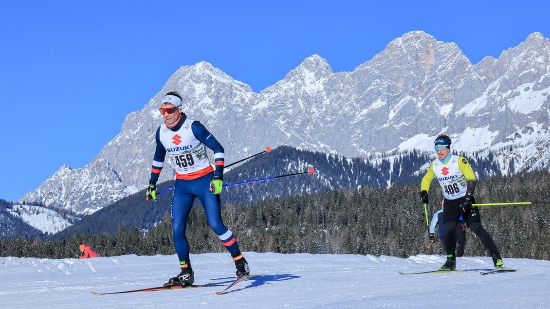 Zwei Skifahrer in Wettkampfkleidung laufen auf einer verschneiten Piste. Im Hintergrund sind beeindruckende Berge und ein klarer blauer Himmel zu sehen. | © Michael Simonlehner