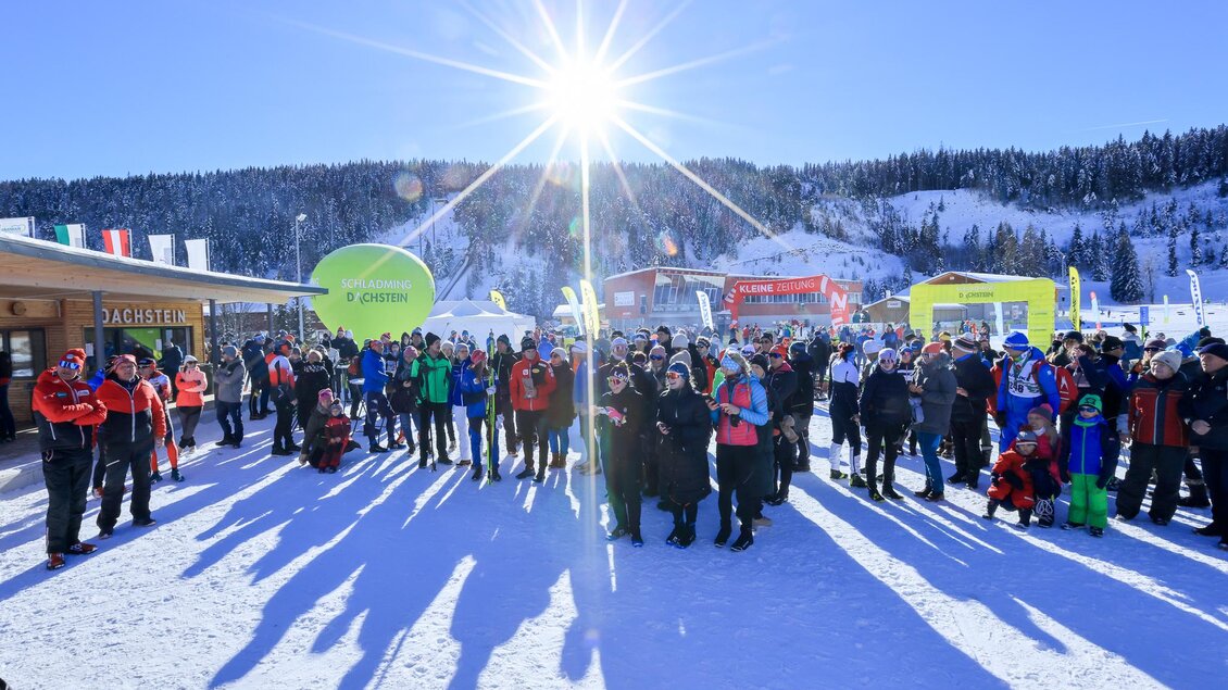 Eine große Menschenmenge steht im Schnee unter einem strahlenden Sonnenlicht. Im Hintergrund sind Berge und Skiausrüstung zu sehen. | © Michael Simonlehner