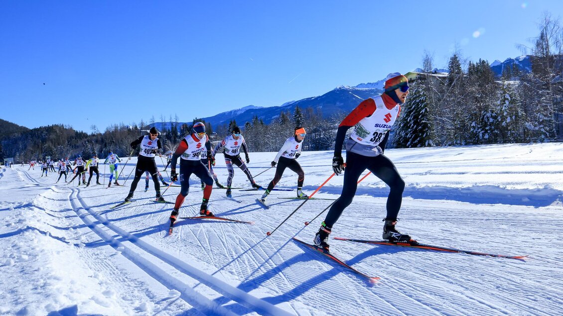 Eine Gruppe von Langläufern bei einem Wettkampf auf einer verschneiten Strecke. Im Hintergrund sind Berge und ein blauer Himmel zu sehen. | © Michael Simonlehner