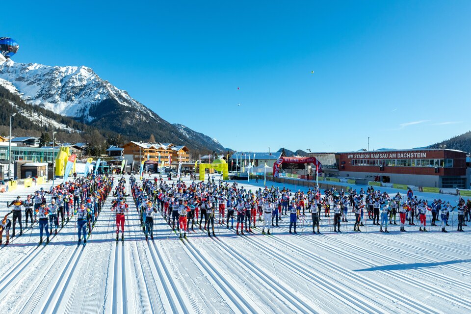 Eine große Gruppe von Teilnehmern steht bereit zum Skilanglauf in einer verschneiten Landschaft. Die Sonne scheint und die Berge sind im Hintergrund sichtbar. | © Hans-Peter Steiner