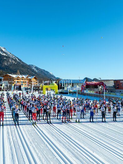 A large group of participants is ready for cross-country skiing in a snowy landscape. The sun is shining and the mountains are visible in the background. | © Hans-Peter Steiner