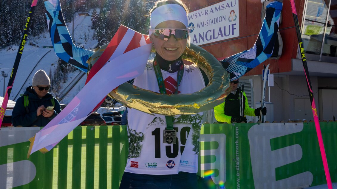 Eine Skilangläuferin hält stolz eine Medaille und eine Flagge in der Hand. Im Hintergrund sind verschneite Berge und ein sonniger Himmel zu sehen. | © Hans-Peter Steiner