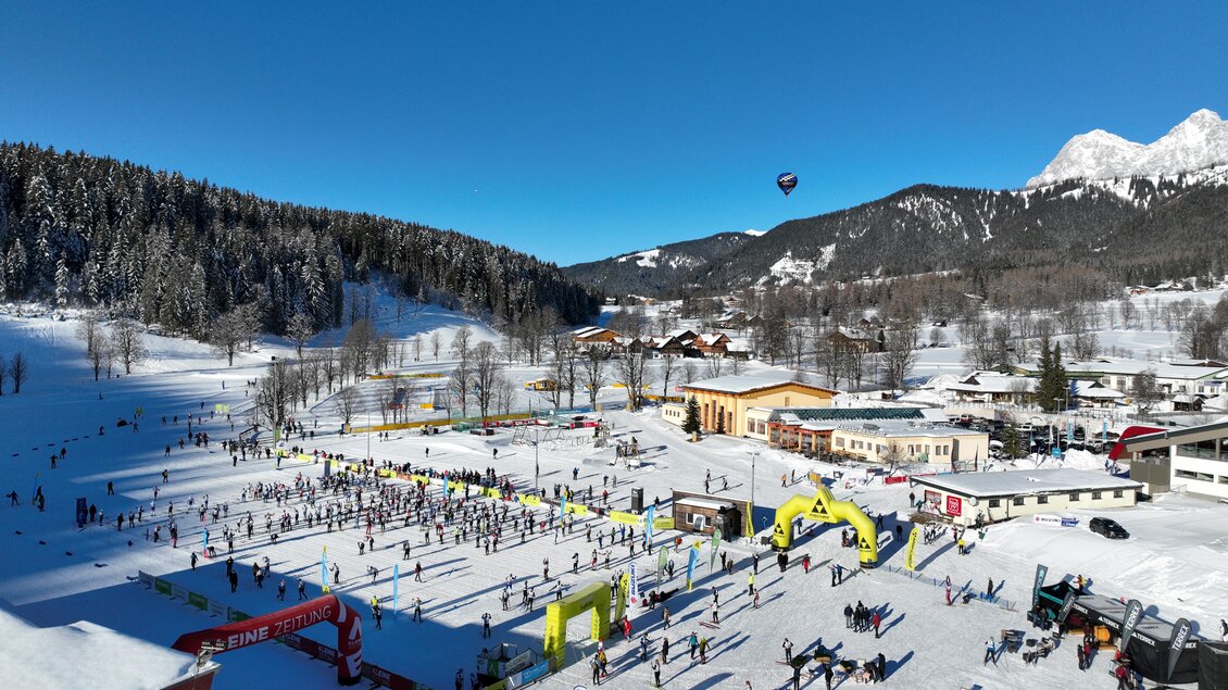 Ein belebter Skihang mit vielen Menschen in einer winterlichen Landschaft. Im Hintergrund sind schneebedeckte Berge und ein strahlend blauer Himmel zu sehen. | © Hans-Peter Steiner