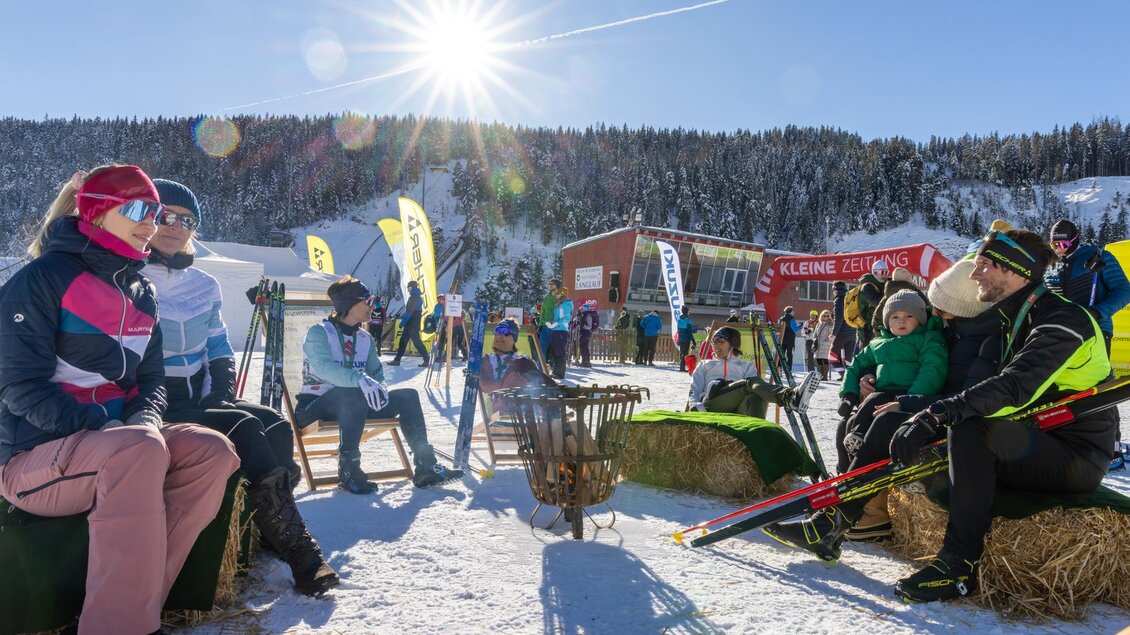 Eine sonnige Winterszenen mit Menschen, die im Schnee entspannen und sich vor einem Lagerfeuer auf Strohballen sitzen. Im Hintergrund sind Skifahrer und eine Berglandschaft zu sehen. | © Hans-Peter Steiner