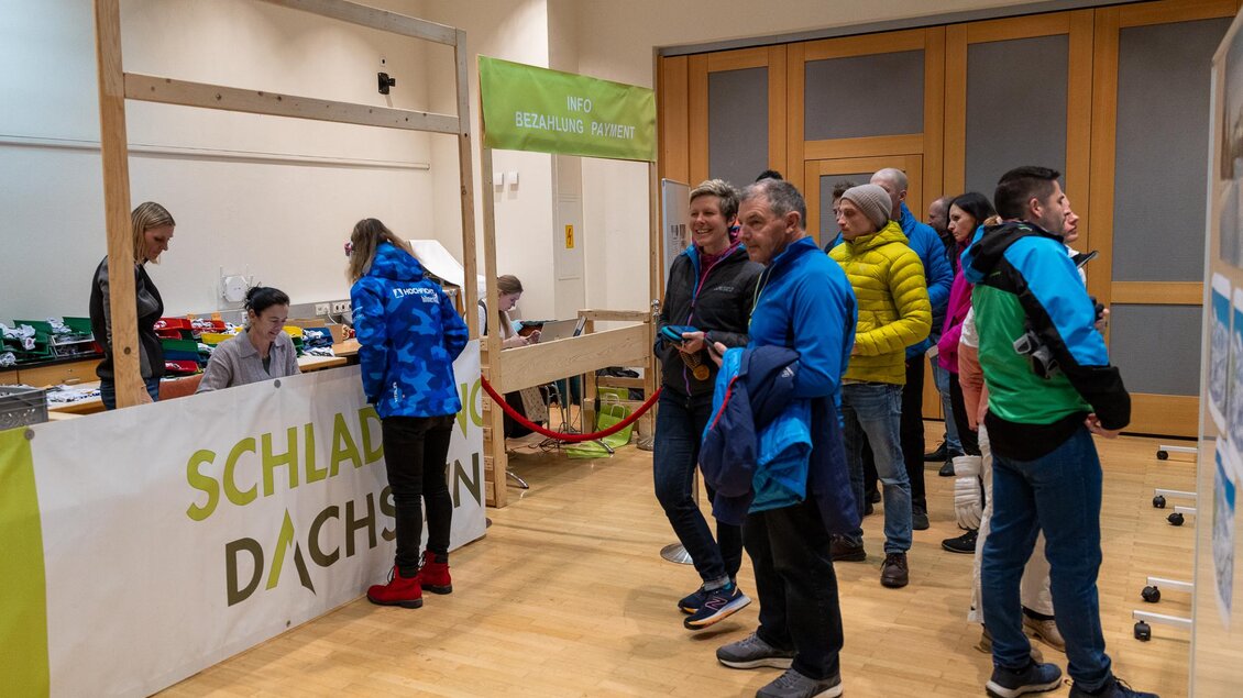 Eine Gruppe von Menschen steht an einem Anmeldestand mit dem Schriftzug „Schladming Dachstein“. Das Umfeld sieht nach einer Veranstaltung oder Messe aus. | © Michael Simonlehner