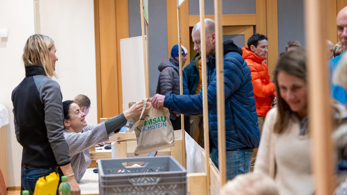Eine Gruppe von Menschen in einem Raum, in dem Waren ausgetauscht werden. Eine Person übergibt eine Tasche an einen anderen, während weitere Personen im Hintergrund warten. | © Michael Simonlehner