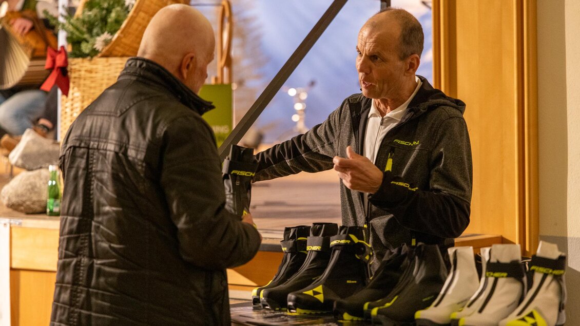 Ein Verkäufer zeigt einem Kunden spezielle Schuhe in einem Verkaufsstand. Im Hintergrund sind dekorative Elemente und weitere Kunden zu sehen. | © Michael Simonlehner