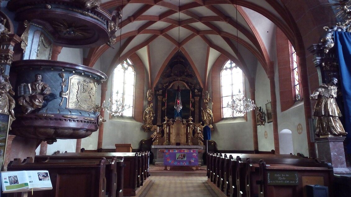 Eine beeindruckende Kircheninneneinrichtung mit einer hohen Decke und schönen Holzverzierungen. Im Hintergrund steht der Altar mit kunstvollen Skulpturen und einer Statue. | © Johanna Provatopoulos