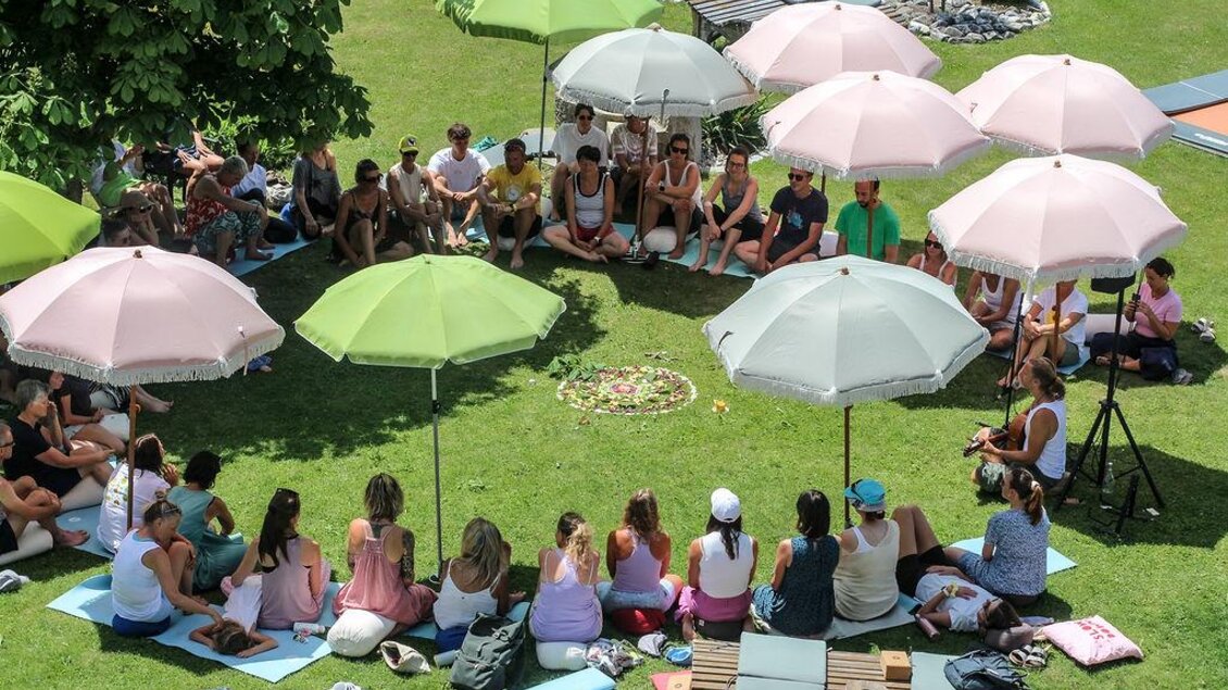 Eine große Gruppe sitzt entspannt im Kreis auf der Wiese. Unter sanften Pastell-Sonnenschirmen entsteht eine ruhige, achtsame Atmosphäre – ideal für Meditation, Austausch und Ankommen. | © Das Bergyogafestival