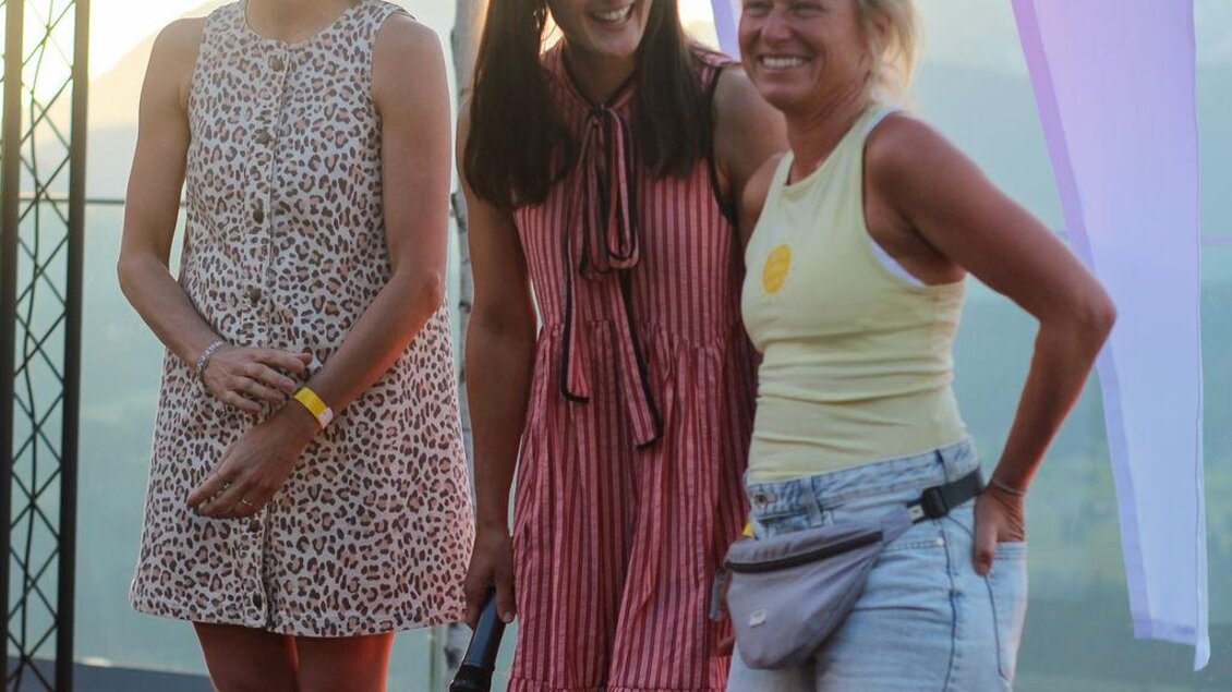 Drei lachende Frauen stehen auf der Bühne vor den Festivalflaggen. Ein stimmungsvoller Moment während eines Programmpunkts beim Bergyoga Festival. | © Das Bergyogafestival