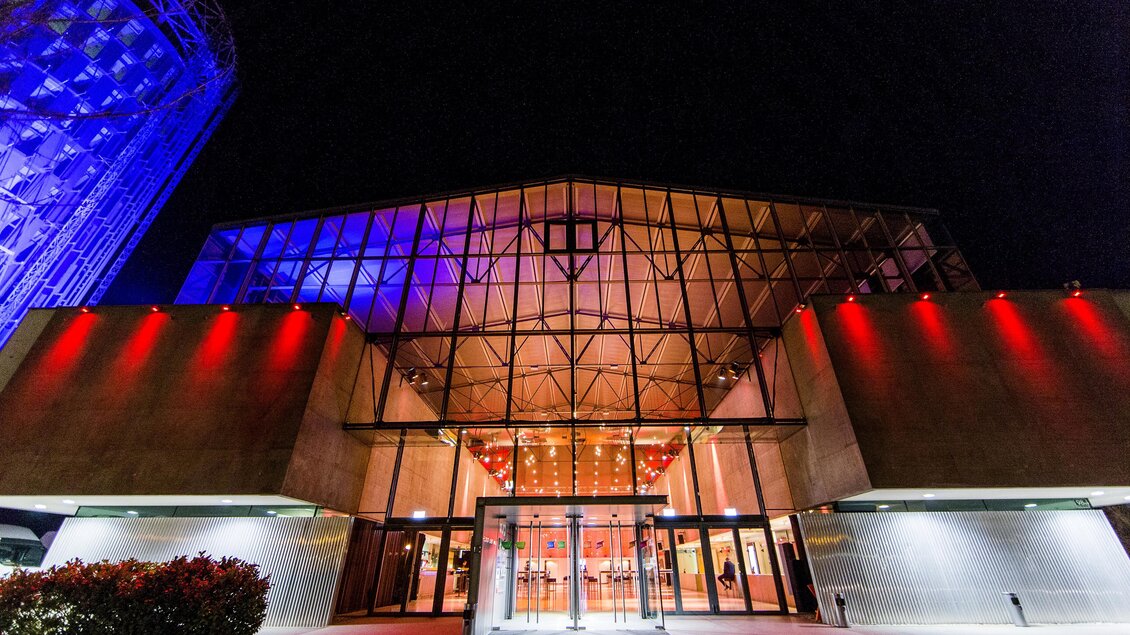 Ein modernes Gebäude bei Nacht mit farbigen Lichtern. Die Glasfront und die architektonischen Details sind gut sichtbar.