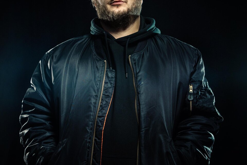 Ein Mensch steht selbstbewusst im Vordergrund, gekleidet in eine dunkle Bomberjacke. Der Hintergrund ist schwarz und hebt die Person hervor. | © ©Antonia Maye