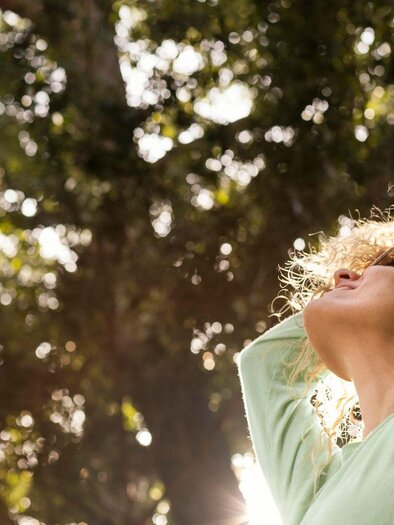 Eine Frau mit lockigem Haar steht unter Bäumen und genießt die Sonne. Sie trägt ein helles Oberteil und hat die Arme behindt.