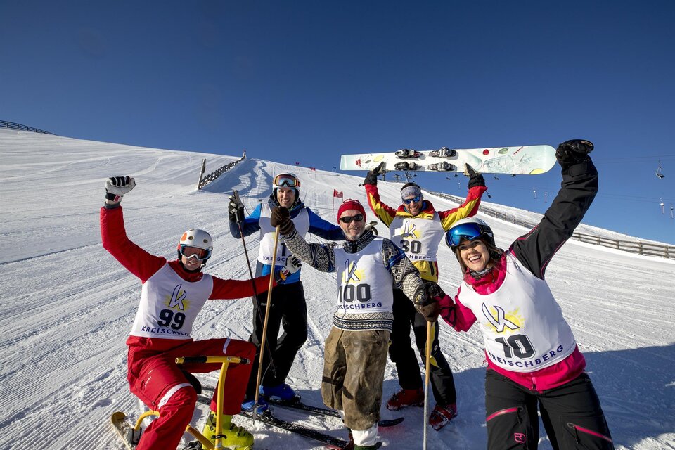 Fünf Skifahrer posieren fröhlich auf einer verschneiten Piste. Sie tragen Wettkampfnummern und haben Spaß bei strahlendem Sonnenschein.