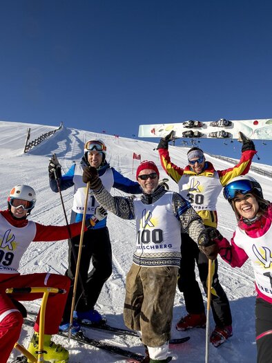 Five skiers are happily posing on a snowy slope. They are wearing competition numbers and having fun in bright sunshine.