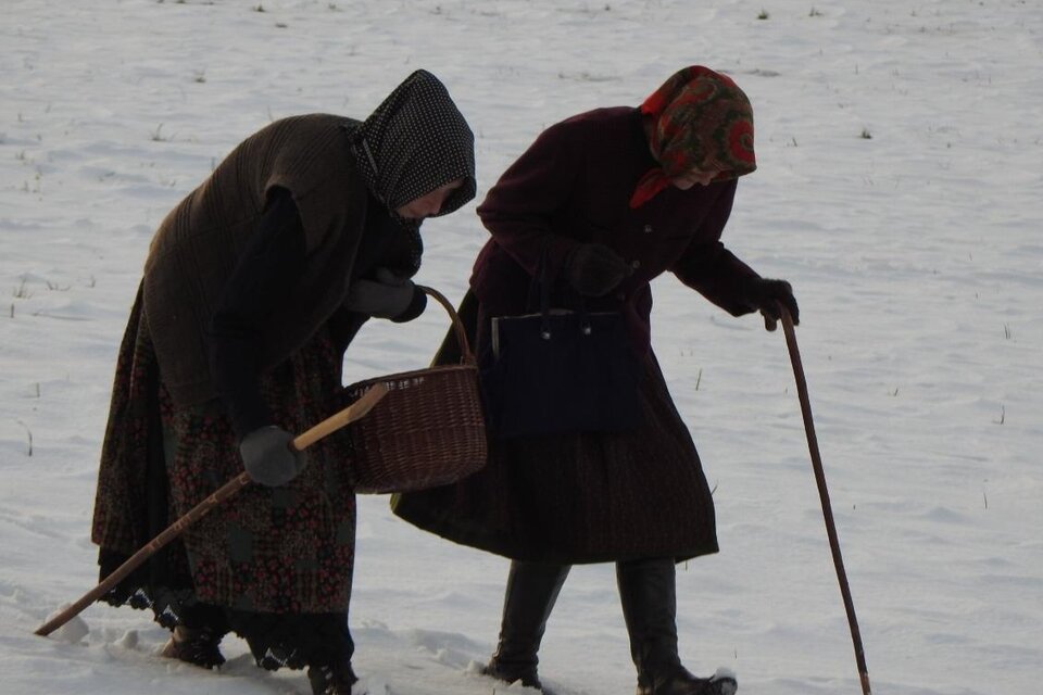 Zwei Frauen in traditionellen Kleidern gehen über einen schneebedeckten Feldweg. Eine der Frauen hält einen Korb und beide haben Stöcke zum Gehen. | © Oststeiermark Tourismus