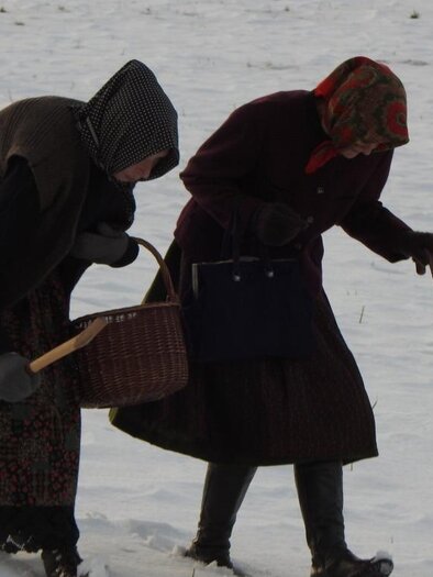 Zwei Frauen in traditionellen Kleidern gehen über einen schneebedeckten Feldweg. Eine der Frauen hält einen Korb und beide haben Stöcke zum Gehen. | © Oststeiermark Tourismus