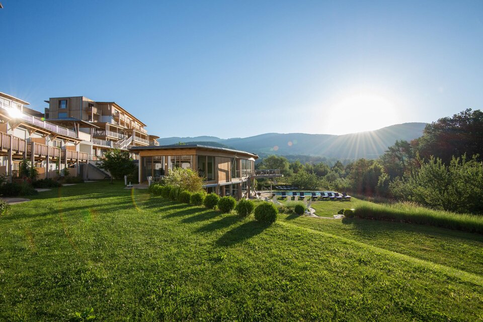 Eine ruhige Landschaft mit einem modernen Gebäude und sanften Hügeln im Hintergrund. Die Sonne geht auf und beleuchtet den grünen Rasen. | © Hotel Retter