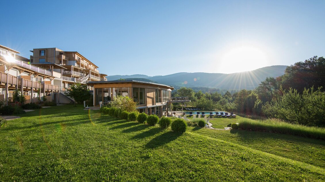 Eine ruhige Landschaft mit einem modernen Gebäude und sanften Hügeln im Hintergrund. Die Sonne geht auf und beleuchtet den grünen Rasen. | © Hotel Retter
