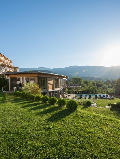 A tranquil landscape with a modern building and gentle hills in the background. The sun is rising and illuminating the green lawn. | © Hotel Retter