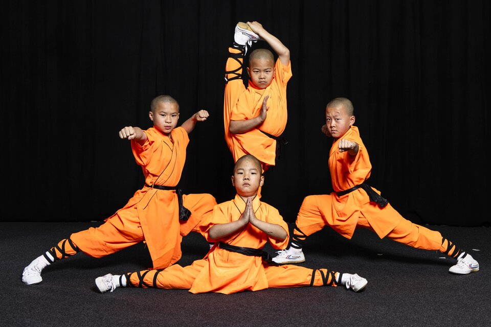 Vier Kinder in orangefarbener Kampfkunstkleidung zeigen beeindruckende Bewegungen. Sie stehen in verschiedenen Posen vor einem schwarzen Hintergrund. | © Die Mönche des Shaolin Kung Fu