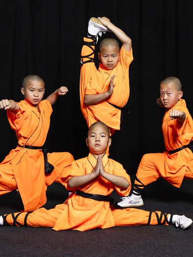Four children in orange martial arts outfits are demonstrating impressive movements. They are standing in various poses against a black background. | © Die Mönche des Shaolin Kung Fu