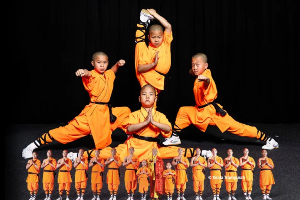 Vier Kinder in orangefarbener Kampfkunstkleidung zeigen beeindruckende Kampfsportposen. Der Hintergrund ist schwarz, wodurch die Bewegungen und die Kleidung hervorgehoben werden.