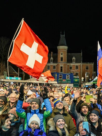 A large crowd is celebrating at night and holding up a Swiss flag. In the background, an impressive building can be seen. | © Martin Huber
