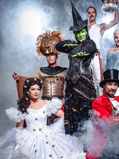 A group of actors in colorful costumes stands on a stage, surrounded by fog. The characters include a witch, a princess, and other imaginative figures. | © PURA VIDA THEATER UND MUSICAL GMBH