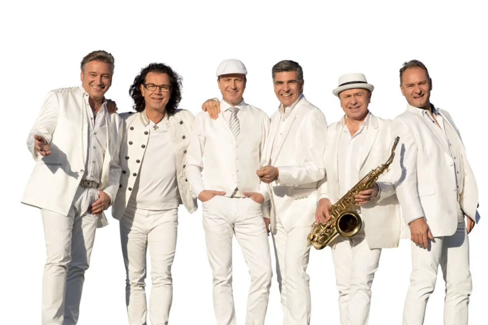Eine Gruppe von sechs Herren in weißen Anzügen steht zusammen. Sie lächeln freundlich und halten verschiedene Musikinstrumente, darunter ein Saxophon. | © Die Paldauer