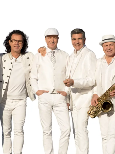 A group of six gentlemen in white suits stands together. They are smiling warmly and holding various musical instruments, including a saxophone. | © Die Paldauer