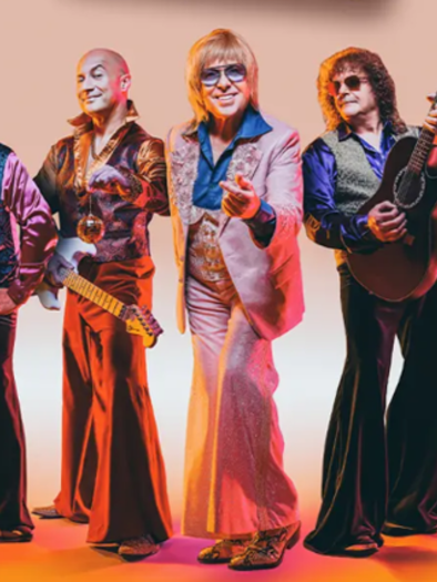 A group of nine musicians in colorful, vintage outfits stands together. They hold various musical instruments and smile at the camera. | © Oststeiermark Tourismus