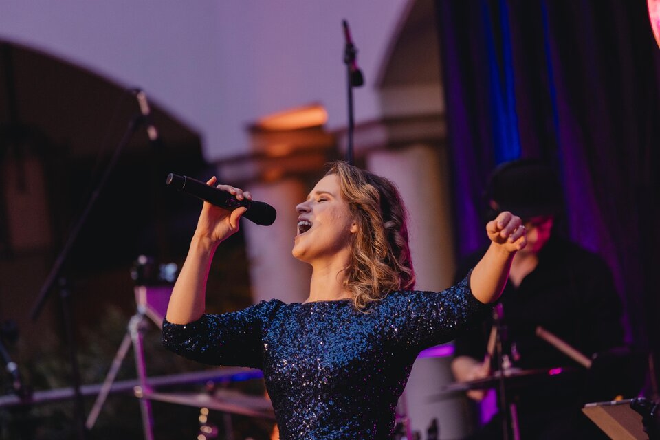 Eine Sängerin mit einem Mikrofon in der Hand tritt auf einer Bühne auf. Im Hintergrund sind Musiker und Bühnenlichter zu sehen. | © Karl Schrotter