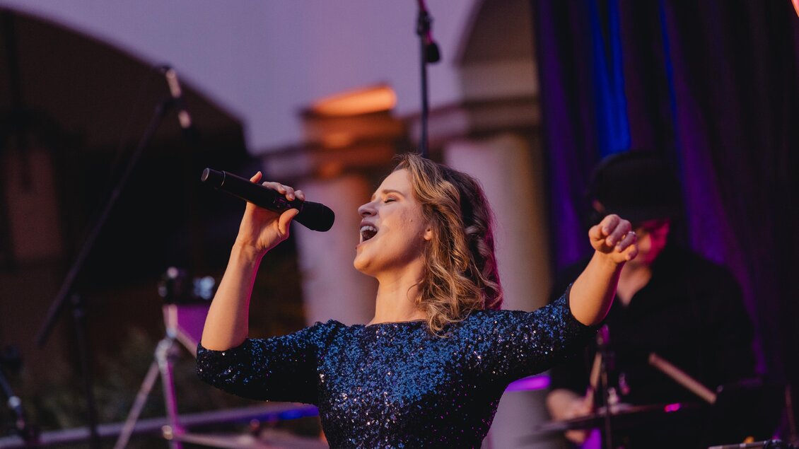 Eine Sängerin mit einem Mikrofon in der Hand tritt auf einer Bühne auf. Im Hintergrund sind Musiker und Bühnenlichter zu sehen. | © Karl Schrotter