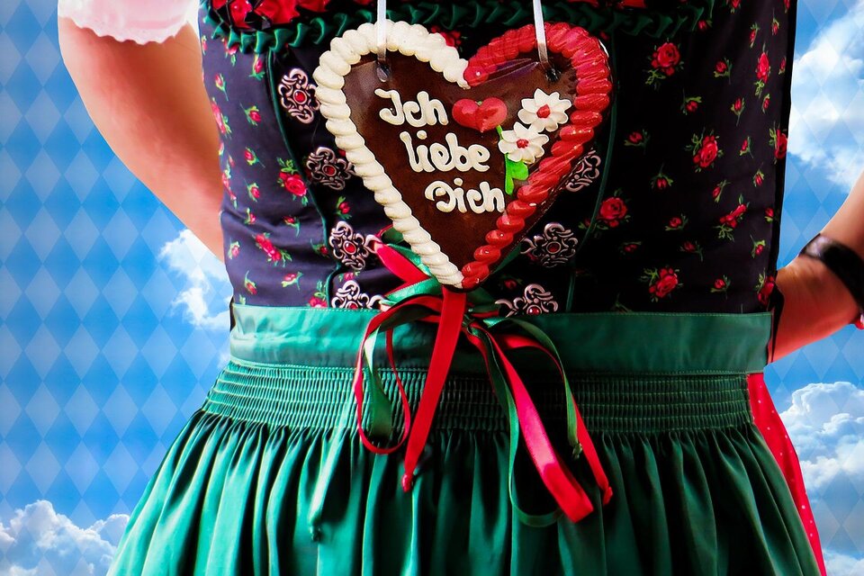 Eine Person trägt ein traditionelles Dirndl mit einer großen Lebkuchenherz-Plakette, auf der "Ich liebe Dich" steht. Der Hintergrund zeigt einen klaren Himmel mit Wolken. | © Symbolfoto Pixabay