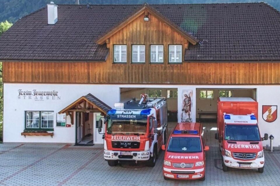 Ein Feuerwehrhaus mit Holzverkleidung und drei Einsatzfahrzeugen davor. Im Hintergrund sind Berge zu sehen. | © Rüsthaus der Freiwiligen Feuerwehr Strassen