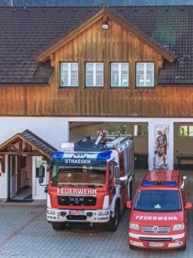 A fire station with wooden cladding and three emergency vehicles in front. In the background, mountains can be seen. | © Rüsthaus der Freiwiligen Feuerwehr Strassen