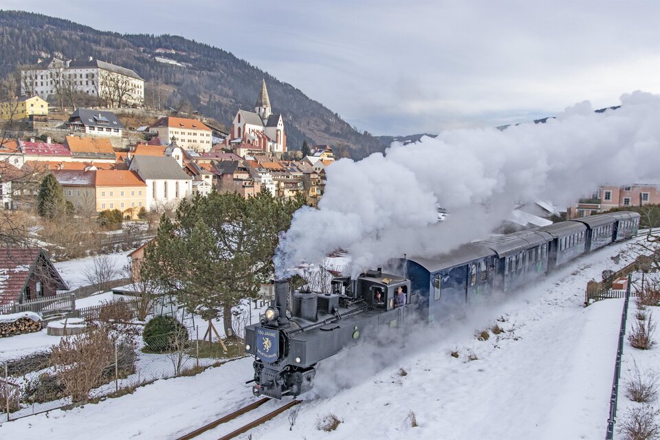 Eine Dampflok fährt durch eine verschneite Landschaft mit einem kleinen Dorf im Hintergrund. Die Szene zeigt eine malerische Winterlandschaft mit Schnee und Dampf. | © Tv Murau