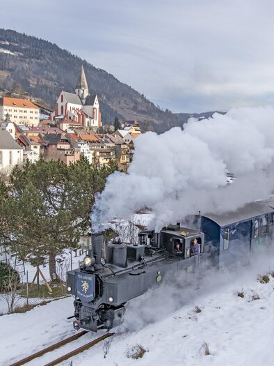 A steam locomotive travels through a snowy landscape with a small village in the background. The scene depicts a picturesque winter landscape with snow and steam. | © Tv Murau
