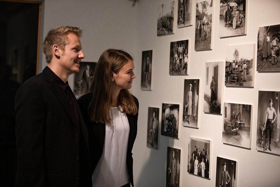 Ein Paar betrachtet historische Schwarz-Weiß-Fotografien an einer Wand. Die Bilder zeigen verschiedene Menschen und Szenen aus der Vergangenheit. | © TV Südsteiermark - nadinegeuterphotography