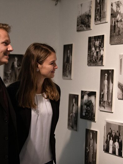 A couple is looking at historical black-and-white photographs on a wall. The images depict various people and scenes from the past. | © TV Südsteiermark - nadinegeuterphotography