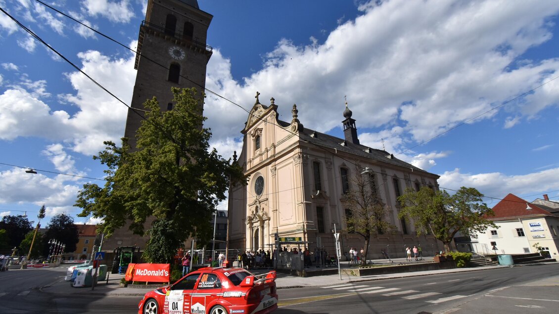 Ein rotes Auto fährt auf einer Straße neben einer historischen Kirche mit einem hohen Turm. Der Himmel ist klar und hat einige Wolken. | © Peter Hopf