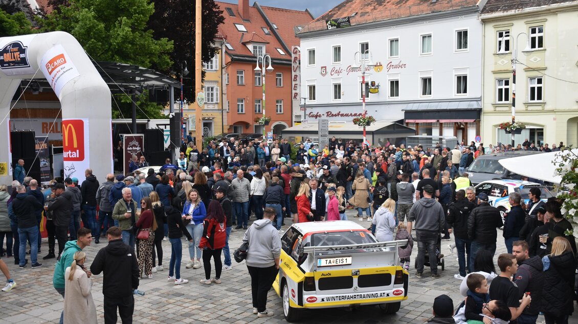 Eine belebte Stadtplatz mit vielen Menschen. Im Vordergrund steht ein auffälliges Rennfahrzeug. | © Peter Hopf