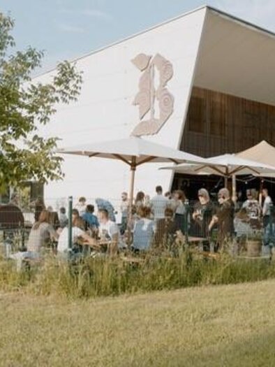 A modern café with an outdoor area and many guests. The space is surrounded by a green meadow and trees. | © Brauhaus Bevog